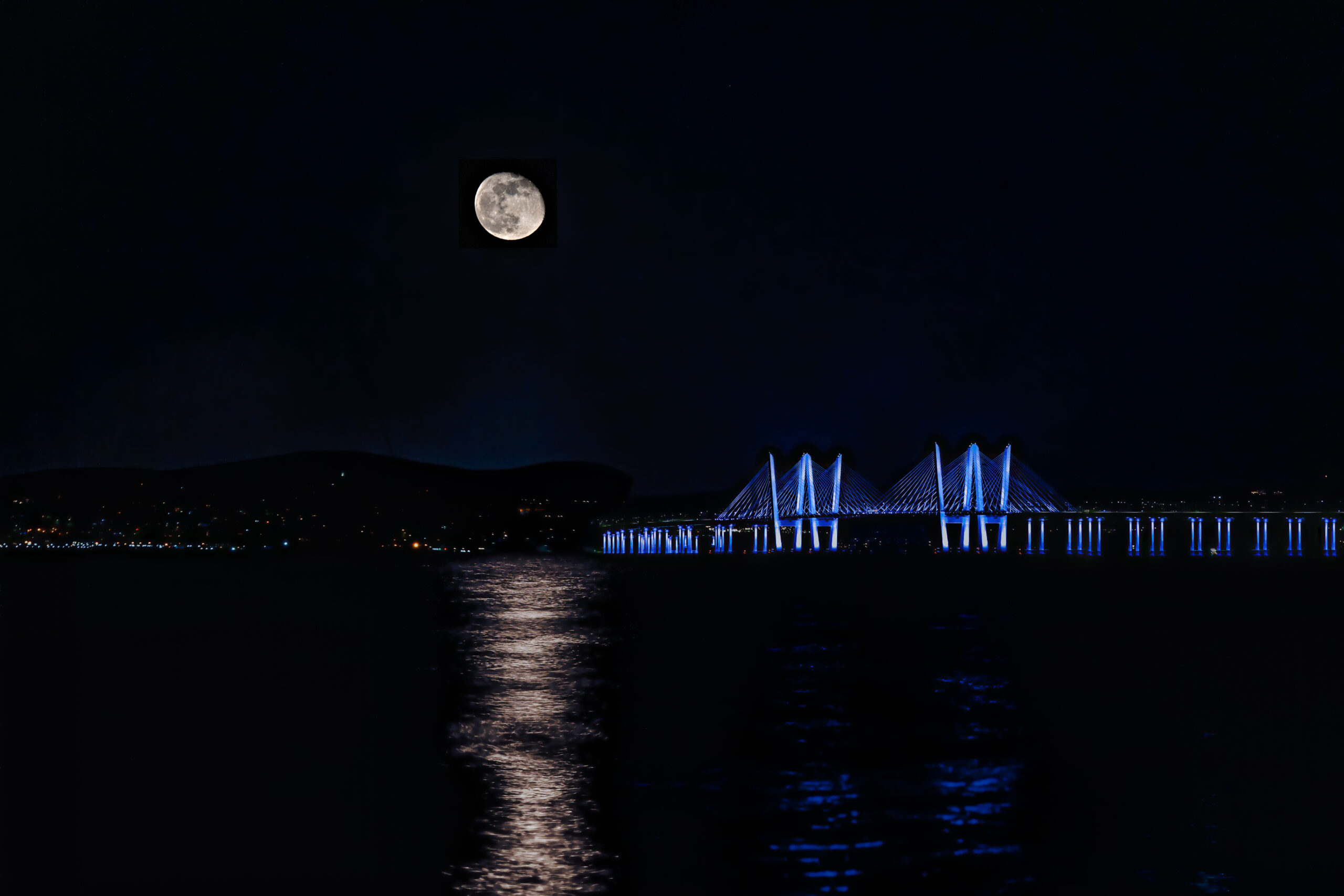 Almost full moon rising over river and bridge lit with blue lights.