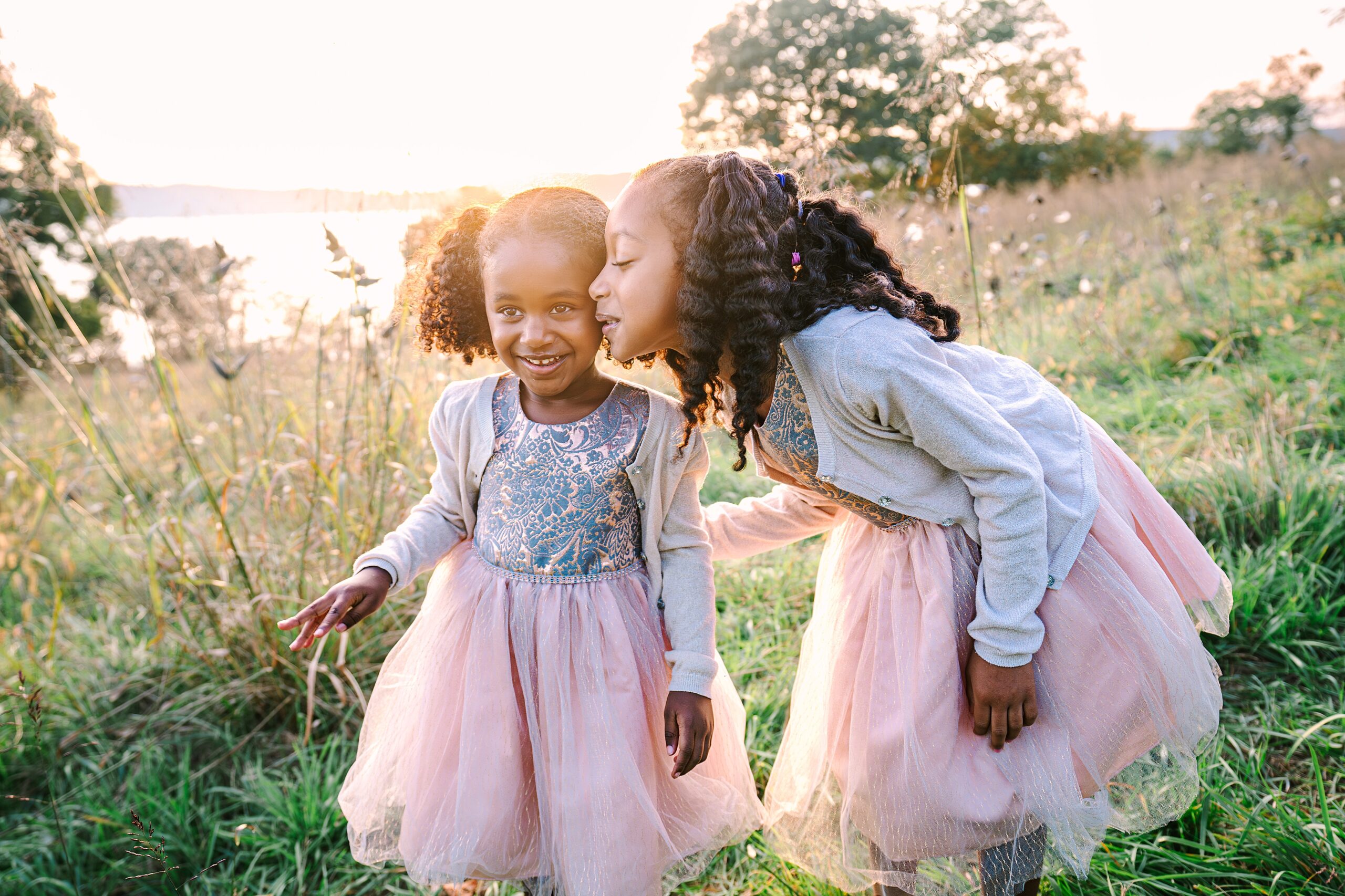 Two young sisters in pink dresses laughing in an open field.