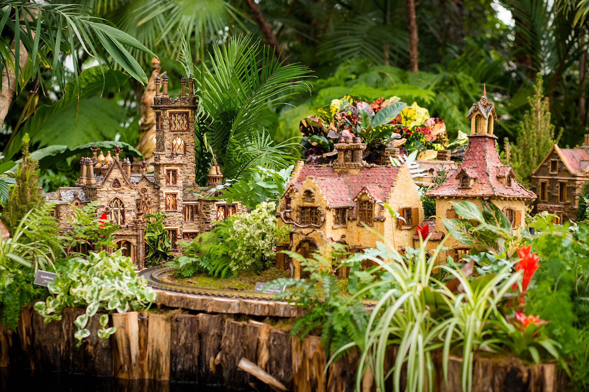 Miniature model village displayed among lush plants at NYBG Holiday Train Show.