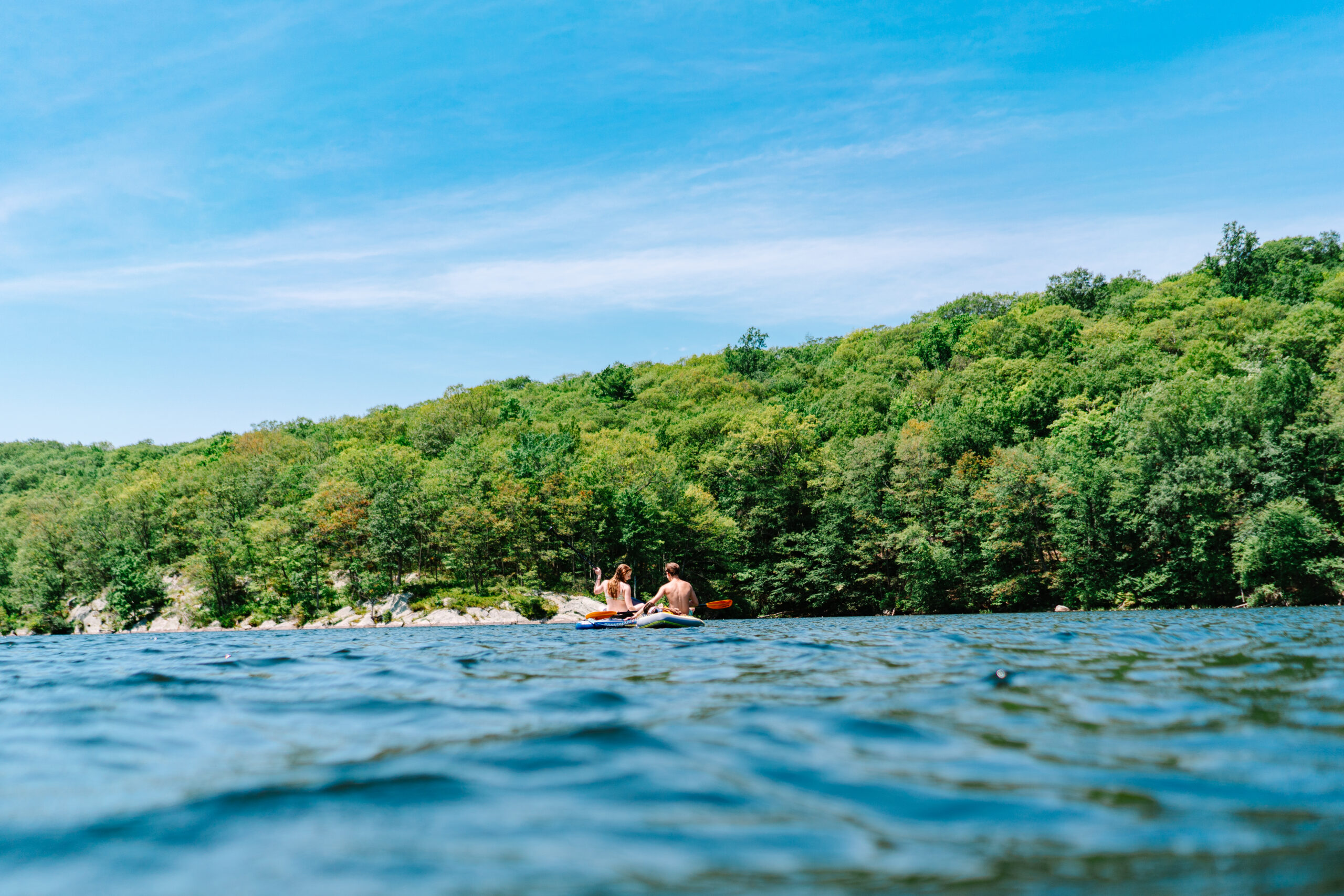Two teenagers paddleboarding on a scenic lake surrounded by forest in the Hudson Valley during summer.